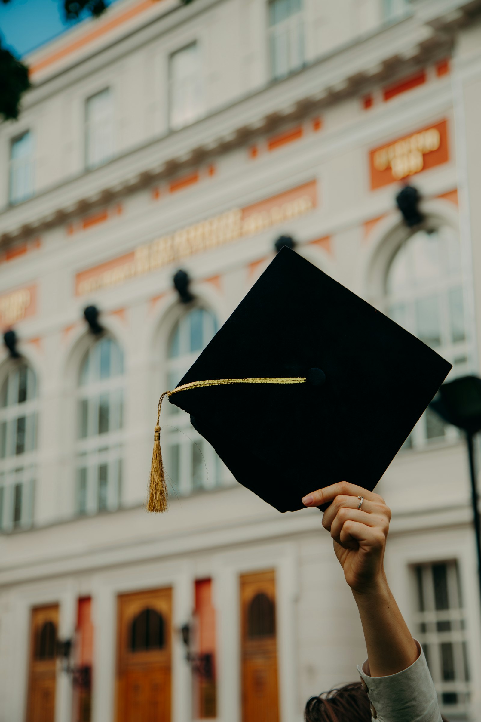 A graduate holding up a cap with a gold tassel in front of a university